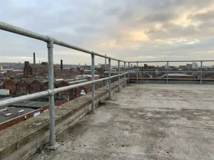 Silbernes Sicherheitsgeländer auf einem Flachdach mit Blick auf eine industrielle Stadtsilhouette bei bedecktem Himmel.
